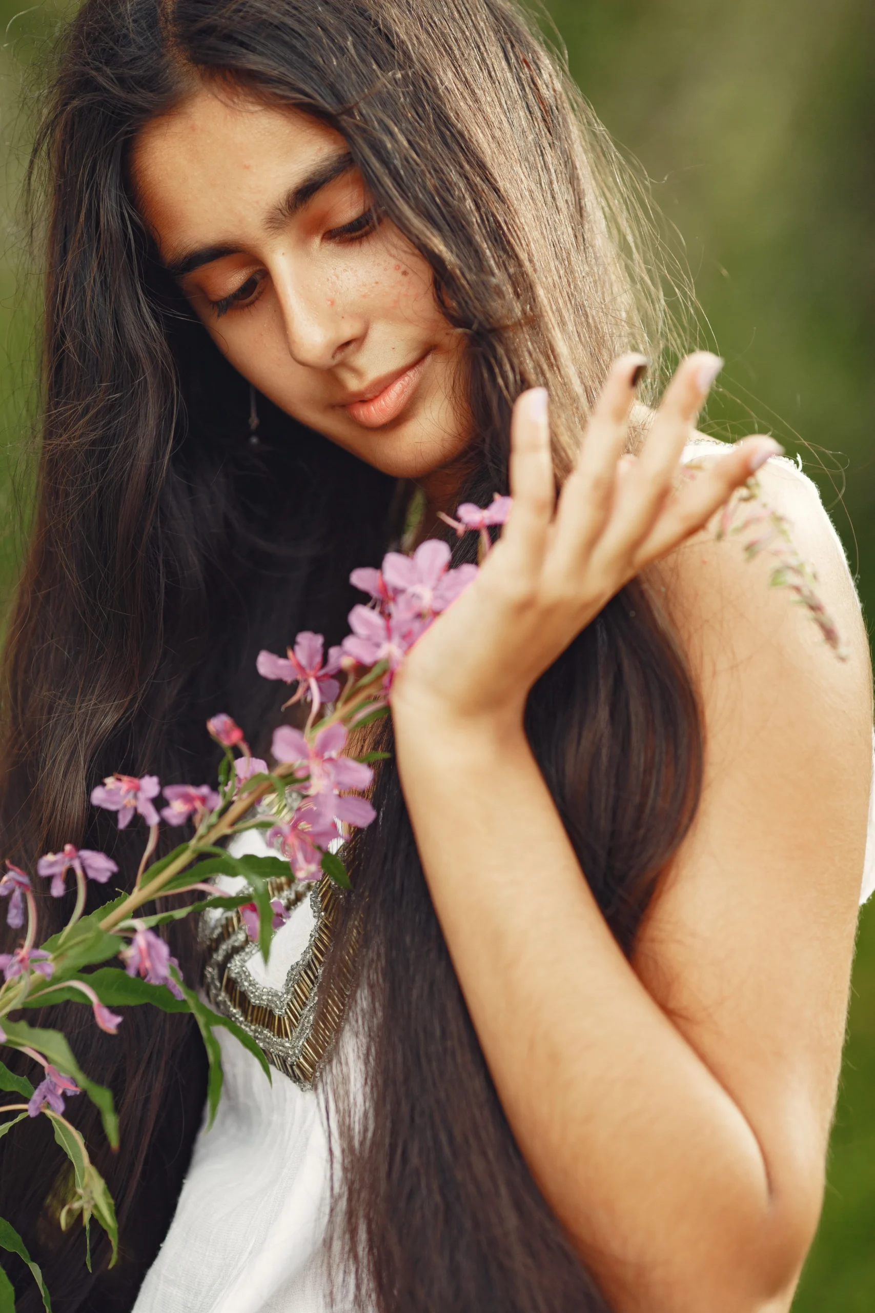 En indisk kvinna med långt, mörkt hår håller varsamt i rosa blommor och ler mjukt i en naturlig miljö. Hon bär en blå klänning omgiven av grönska, som förkroppsligar essensen av indisk-kvinna-med-långt-hår-dam-blå-klänning-flicka-med-orörd-natur.